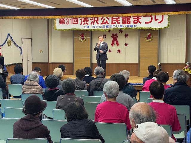 イスに座った多数の出席者の前で立って挨拶する市長