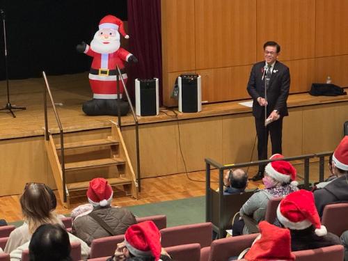 階段状の席に座るサンタ帽をかぶった出席者に立って挨拶をする市長。ステージの上にはサンタクロースの大きな人形が立っている。
