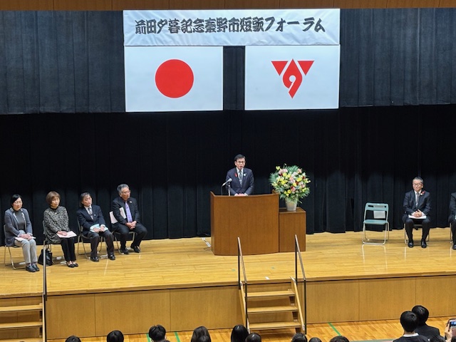 ステージの上で演台の後ろに立って挨拶する市長、左側には来賓が4名座っている。