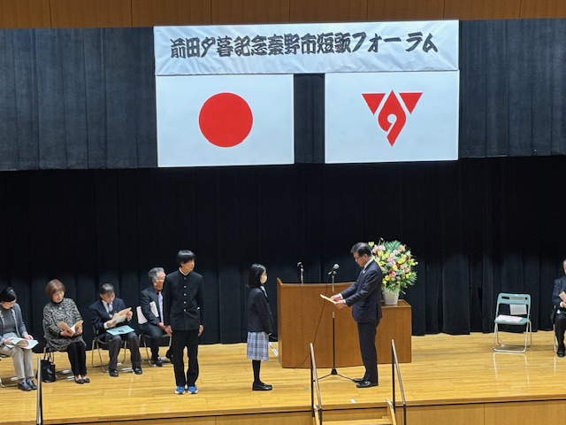 ステージの上で制服姿の中学生の男女が市長から賞状を受け取っている。