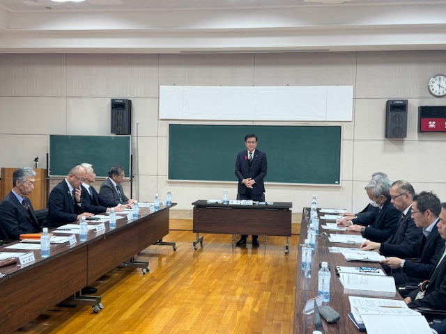 ロの字型の会議室でスーツの男性が会議をしている。市長は黒板の前で立って挨拶をしている。