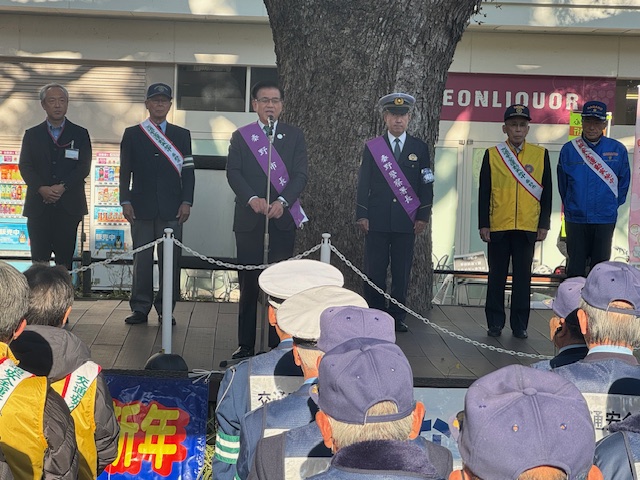 大きなケヤキの木の下で、キャップや制帽をかぶった男性たちの前で挨拶をする市長。