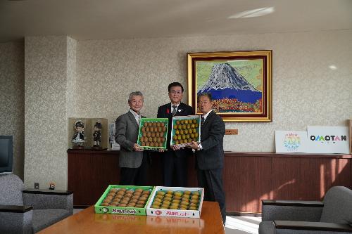 市長を中心に2人のスーツの生産者の男性が箱に詰まったキウイフルーツを持っている。