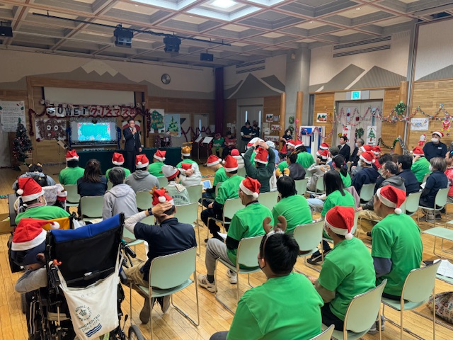 緑のジャンバーにサンタ帽をかぶった大勢の人が市長の挨拶を聞いている