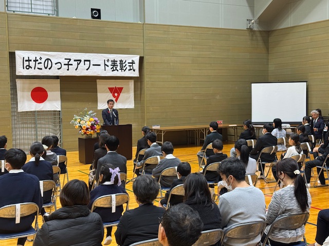 手前に大勢の子供と保護者が椅子に座り、奥に市長が演台の後ろで挨拶をしている。市長の頭上にはだのっ子アワード表彰式の看板がある。