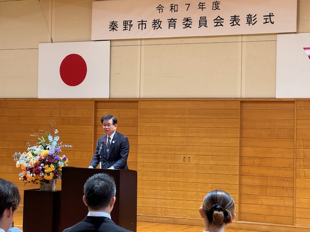 秦野市教育委員会表彰式の看板の下で挨拶をする市長。左側には花が置かれている。
