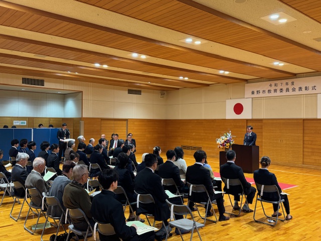 スーツを着て椅子に座る40人ほどの受賞者。奥には市長が演台の後ろで挨拶をしている。