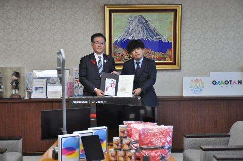 たくさんの家電製品がテーブルの上に並んでいる。奥に感謝状と目録を持った市長とスーツの男性が立っている。