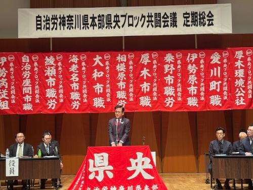 大きな赤い垂れ幕がかかっているステージの前で、大きく県央と書かれた赤い旗が貼られた演台で挨拶する市長。