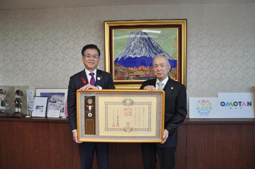 市長とスーツの男性が、賞状と勲章が入った大きな額縁を持って富士山の絵の前で立っている。