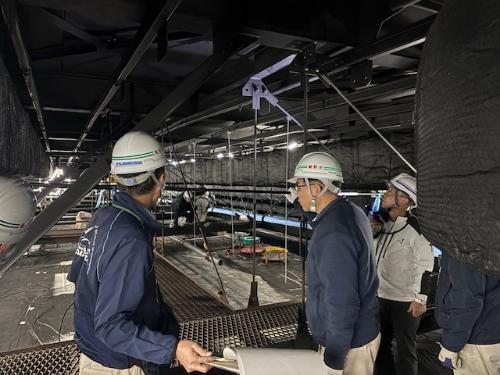 屋根裏の工事現場を見るヘルメットに作業着姿の市長と、説明するヘルメットをかぶった男性