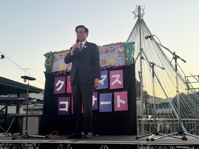 屋外の簡易ステージでマイクを持って挨拶する市長。空は夕暮れ。