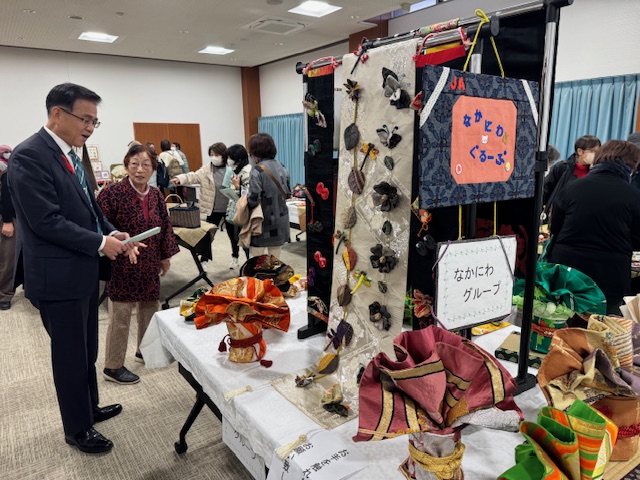 てーぶるの上に展示された手作り作品を鑑賞する市長とそれを説明する女性。