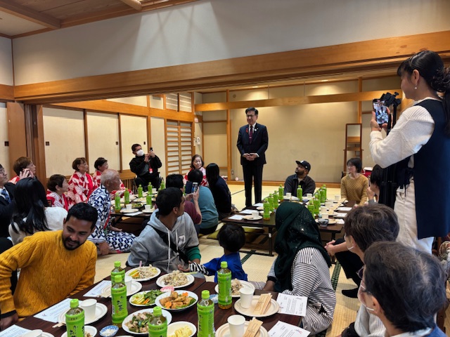 畳の部屋でテーブルに料理が並んでおり、日本人と外国人が一緒に座っている。奥で市長が挨拶をしている。