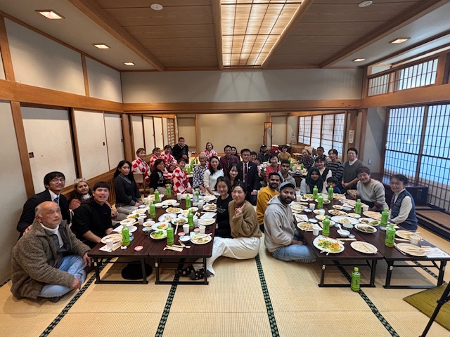 畳敷きの部屋で出席者が40人ほどテーブルを囲み集合写真を撮っている。