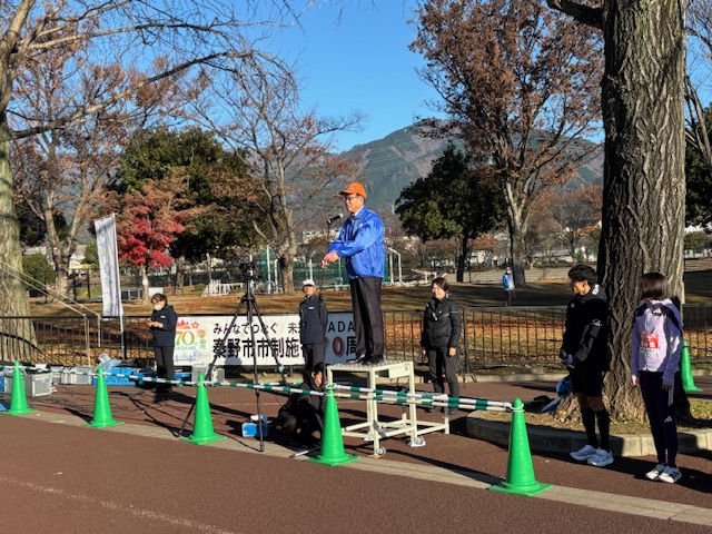 青いジャンバーにオレンジのキャップをかぶった市長が小さな台の上で挨拶をしている。