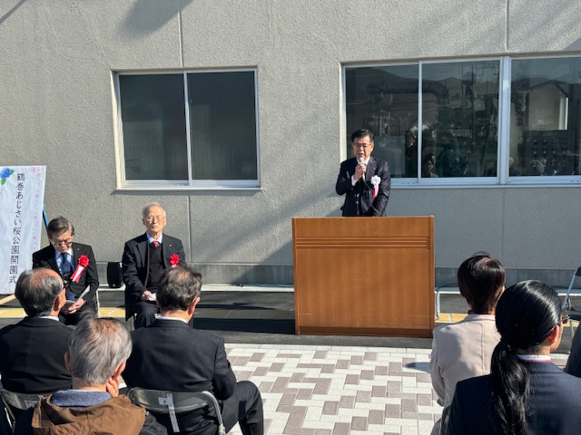 屋外の建物の前で演台に立って挨拶する市長、左には椅子に座った来賓2名、向かいには出席者が座る