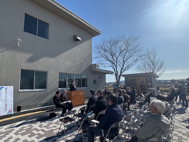 屋外で建物の前に置かれた演台で挨拶する市長と、それを聞く着席した出席者30名ほど。