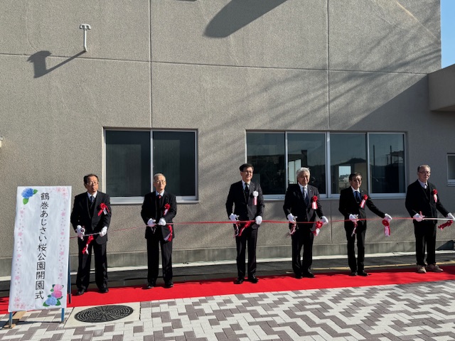 屋外で赤いじゅうたんの上でテープカットをするスーツの男性6人