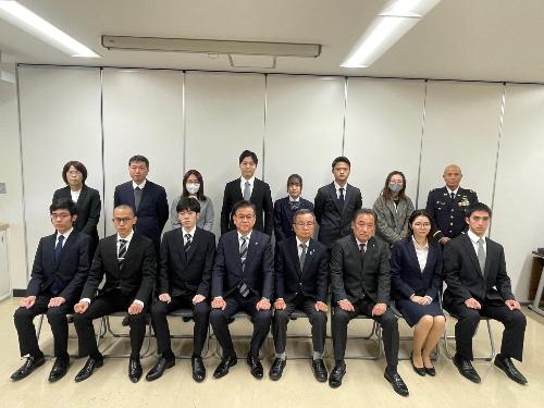 スーツの男女が前列に8人椅子に座り、後列に8人立っている。