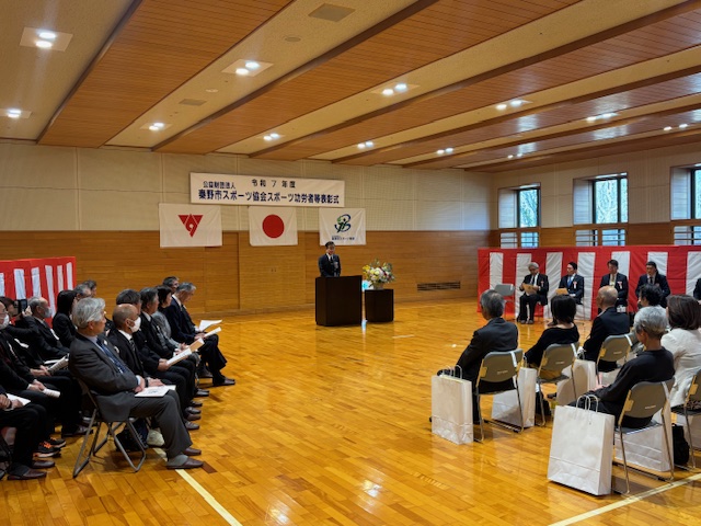 演台に立って挨拶する市長の両側に紅白幕があり、大勢の来賓が座っている。その向かいに受賞者が10名ほど座っている。