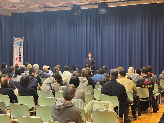 左端に本町公民館まつりののぼり旗が立っている。奥で市長が立って挨拶をし、手前に来場者が多数椅子に座っている。