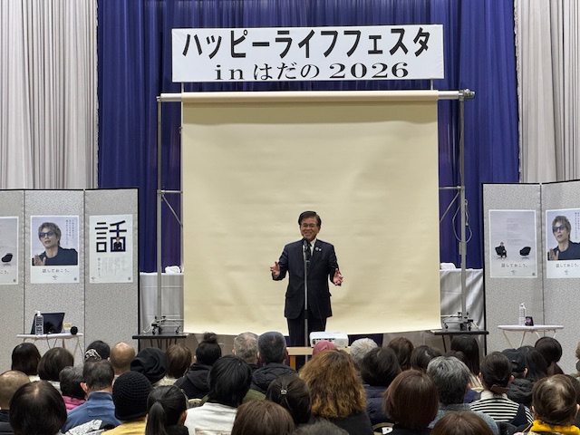 大きなスクリーンの前で立って挨拶をする市長と、手前に大勢の出席者が座っている。