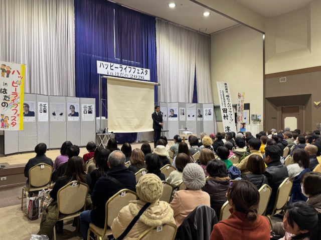 ハッピーライフフェスタの横看板の下で挨拶をする市長の前に100名ほどの出席者が座る。