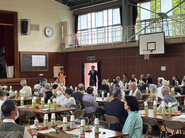 学校の体育館で挨拶する市長とそれを聞く大勢の意高齢者、テーブルにはお茶とお菓子が置いてある