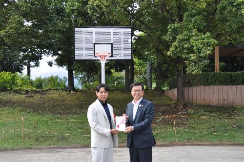 寄贈されたバスケットゴールの前で島半の社長が市長に目録を渡している写真