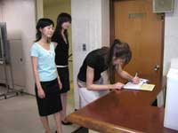 受付カウンターで、黒いシャツの女性が前かがみになって傍聴受付票に書き込んでおり、水色のシャツの女性と黒い服の女性がその後ろに立って見ている様子の写真