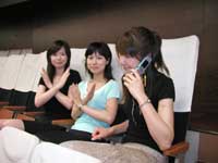 傍聴席に座っている3人の女性のうちの一人が携帯電話で話し、その横の2人の女性が両腕でバツのポーズをとり、携帯電話使用禁止を訴えている様子の写真