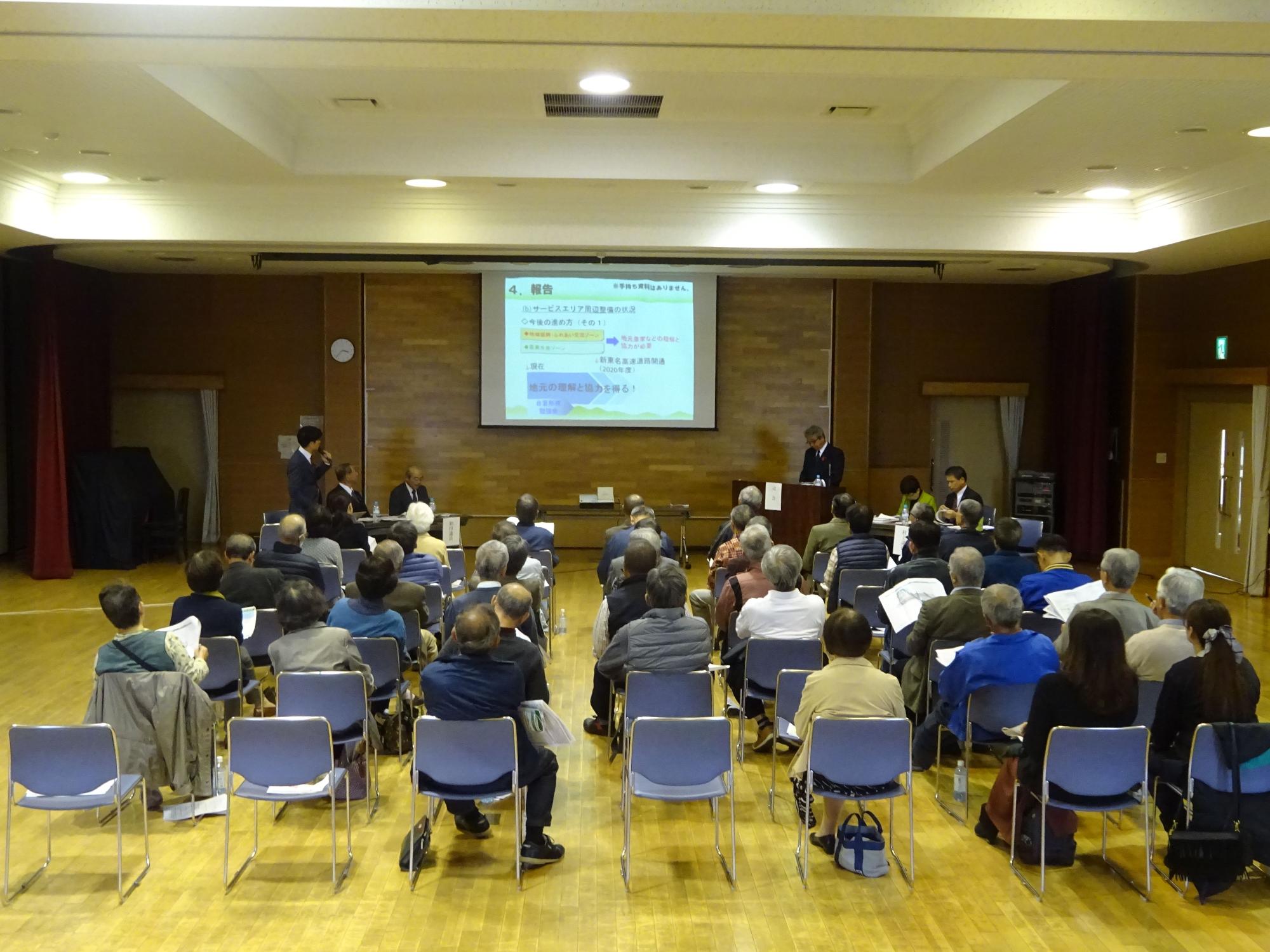 高齢の方々を中心とした多くの人々が椅子に座って前方のスクリーンを見ながら、登壇者による議会報告会が行われている様子の写真