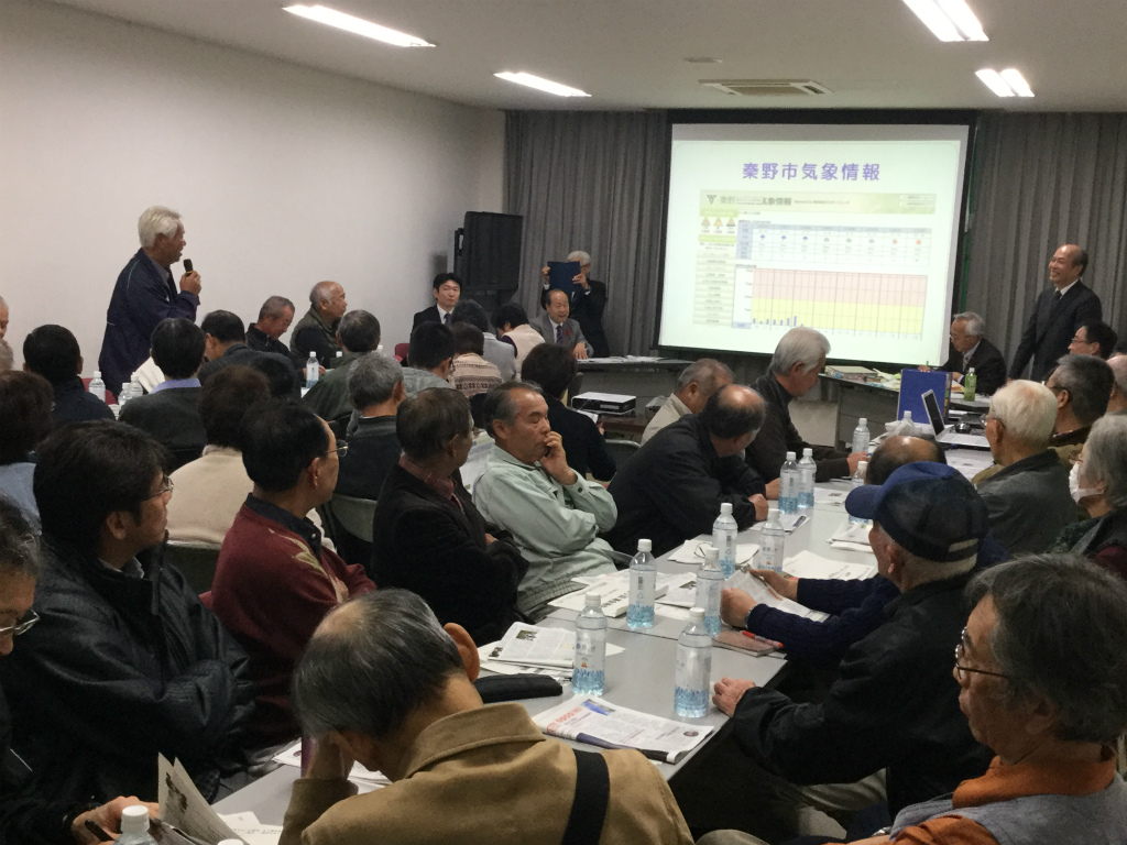 会議室のような場所で、高齢の参加者を中心とした多くの人々がテーブルを囲み、資料が映し出されたスクリーンを見ながら、発言者の話に耳を傾け報告会が行われている様子の写真