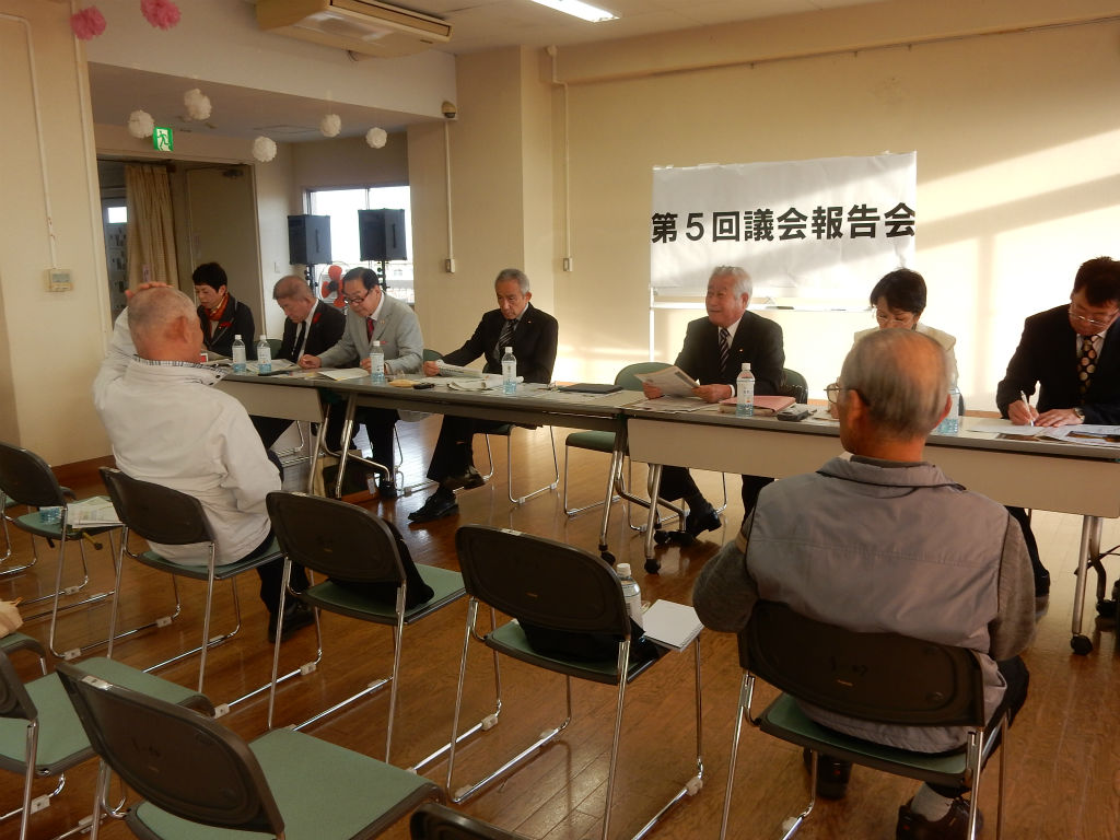 明るい会議室で「第5回議会報告会」と書かれた横断幕の前にスーツ姿の人たちが横一列に座り、数名の住民が向かい合って座って意見交換をしている地域の議会報告会の様子を写した写真