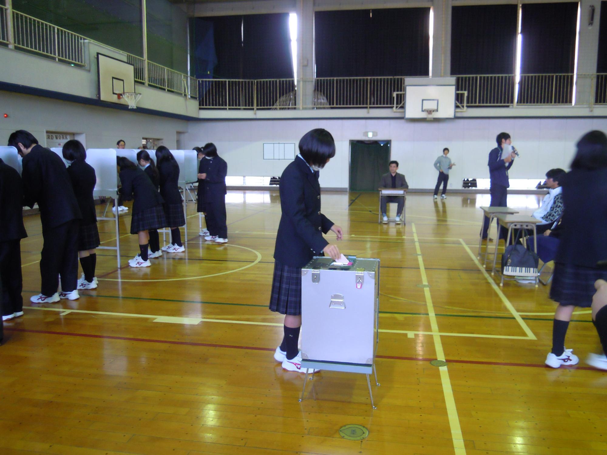 体育館で、制服を着た女子生徒が投票箱に投票用紙を投函しており、他の生徒たちは記載台で用紙の記入や順番待ちをしている模擬投票の風景写真