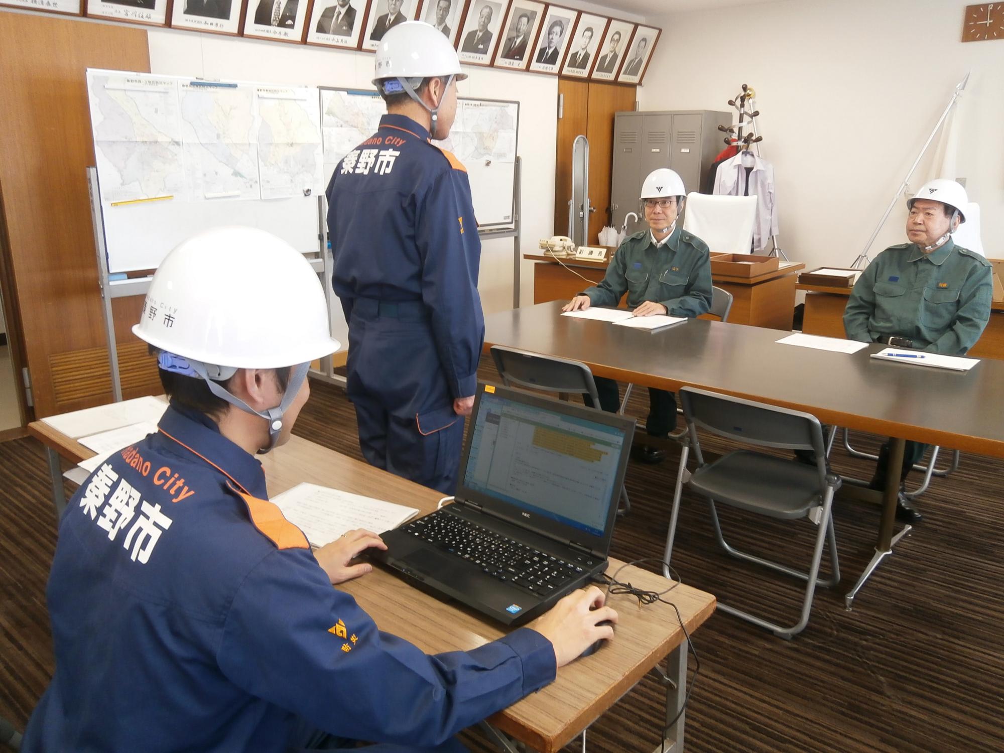 4人の男性が防災服やヘルメットを着用し、消防職員がノートパソコンを操作しつつ、別の消防職員が立って指導をしている安否確認の様子の写真