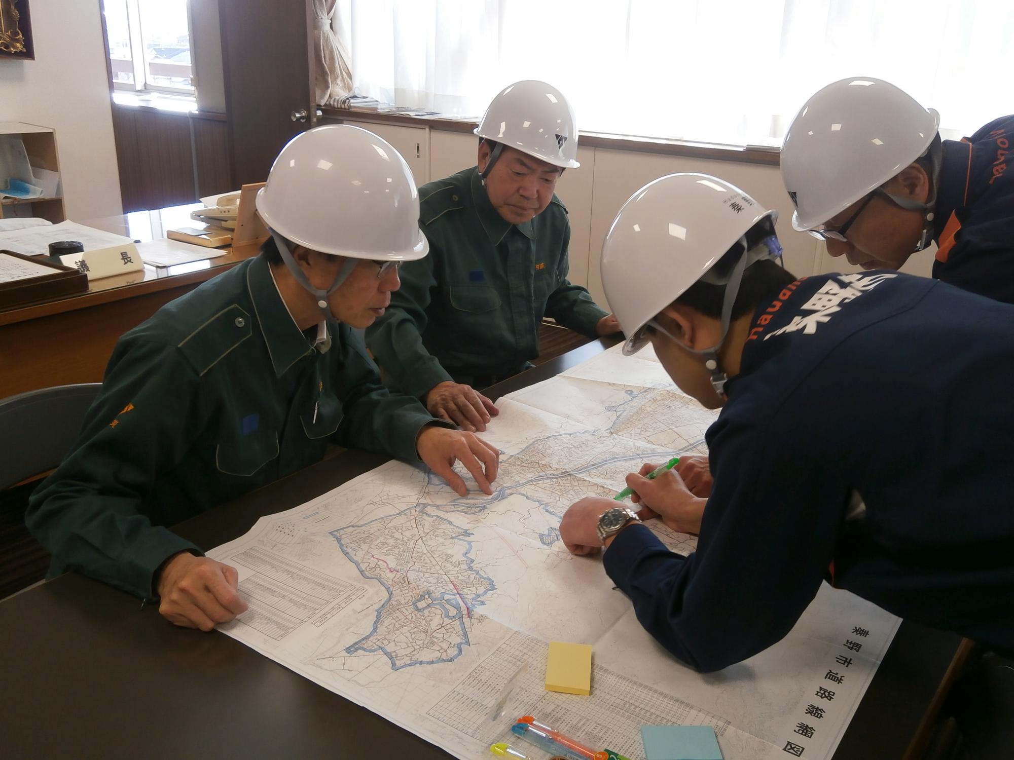 4人の男性がヘルメットを着用し、机の上に広げた地図を囲んで指を差したりペンで書き込んだりしながら、真剣な表情で状況を確認している災害対策会議の様子の写真