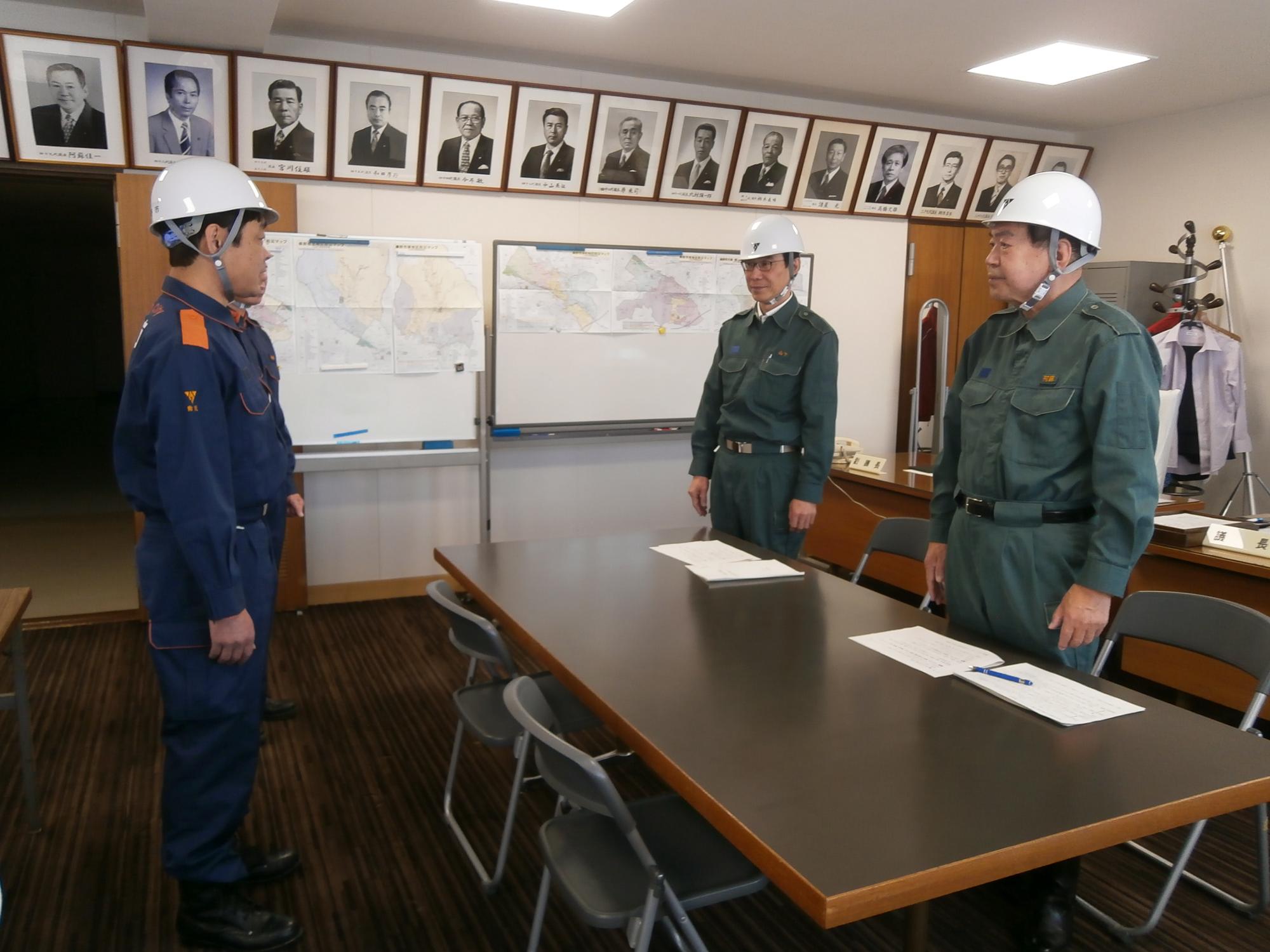 テーブルを挟んで防災服とヘルメットを着用した3人の男性が真剣な面持ちで話し合っている災害対策会議の様子の写真