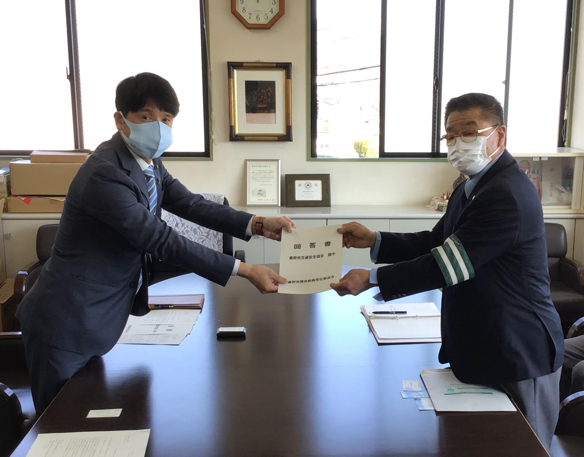 茶色い会議テーブルを挟んで向かい合った、マスクを着用した2人のスーツ姿の男性が、中央で「回答書」の書類を差し出し合う様子の写真