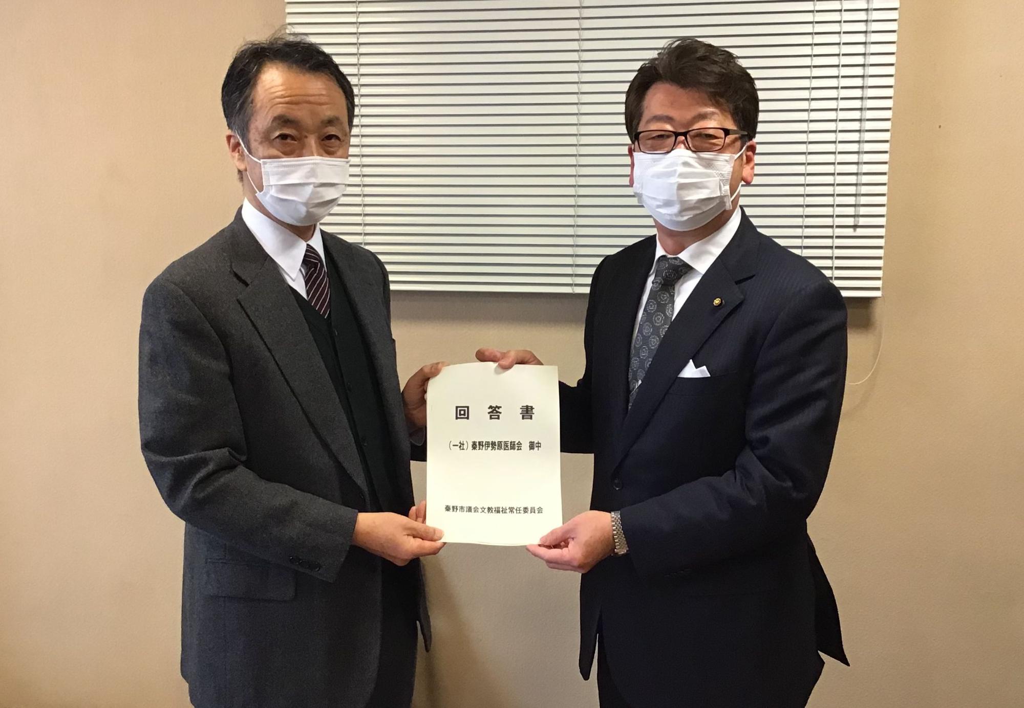 マスクを着用した2人のスーツ姿の男性が中央で「回答書」を両側から持ち、笑顔で受け渡しをしている様子の写真