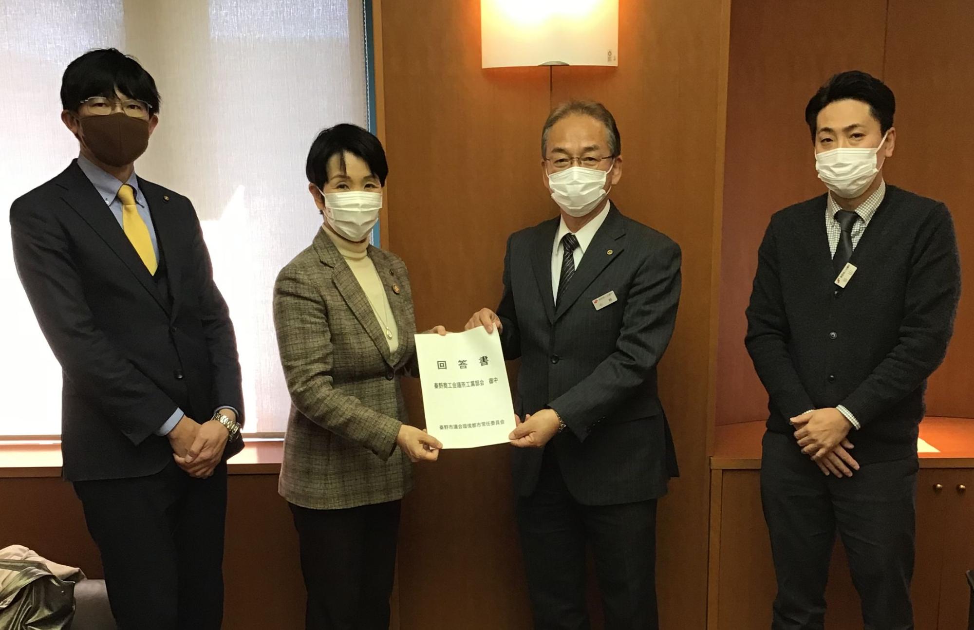 明るい室内にマスクを着用した男女4人が立ち、中央の男女2人が「回答書」と書かれた書類を両側から持ち、カメラに向かって受け渡しをしている様子を捉えた写真