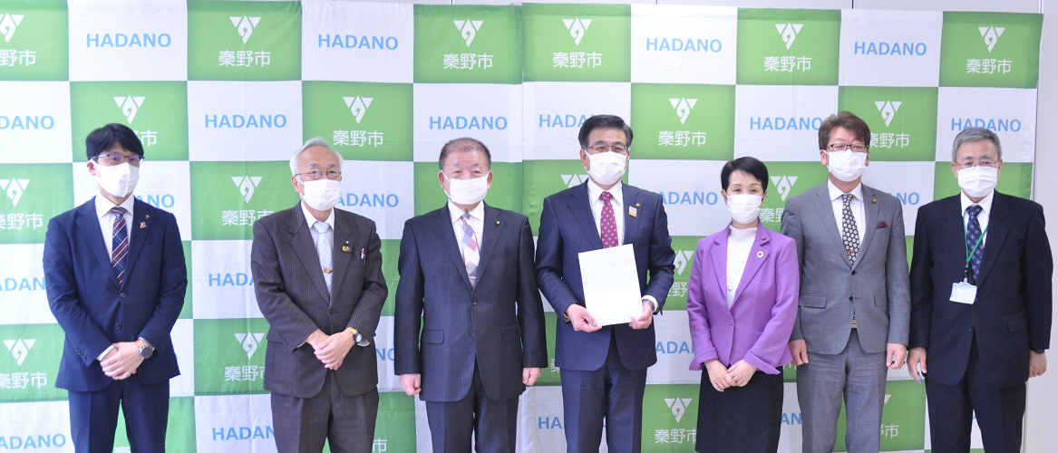 秦野市のバックボードの前で、マスクを着用した市長と男女6人が立ち、中央の市長が要望書を持ち、カメラに向かって並んだ記念写真