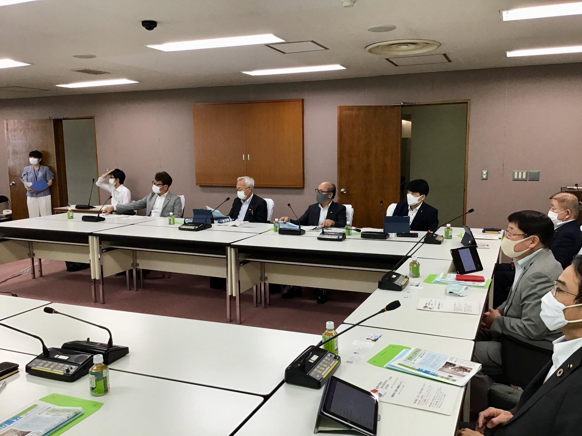 複数の人々がマスクを着用して会議室の長テーブルに座り、タブレットや資料を前にして議論している様子の写真