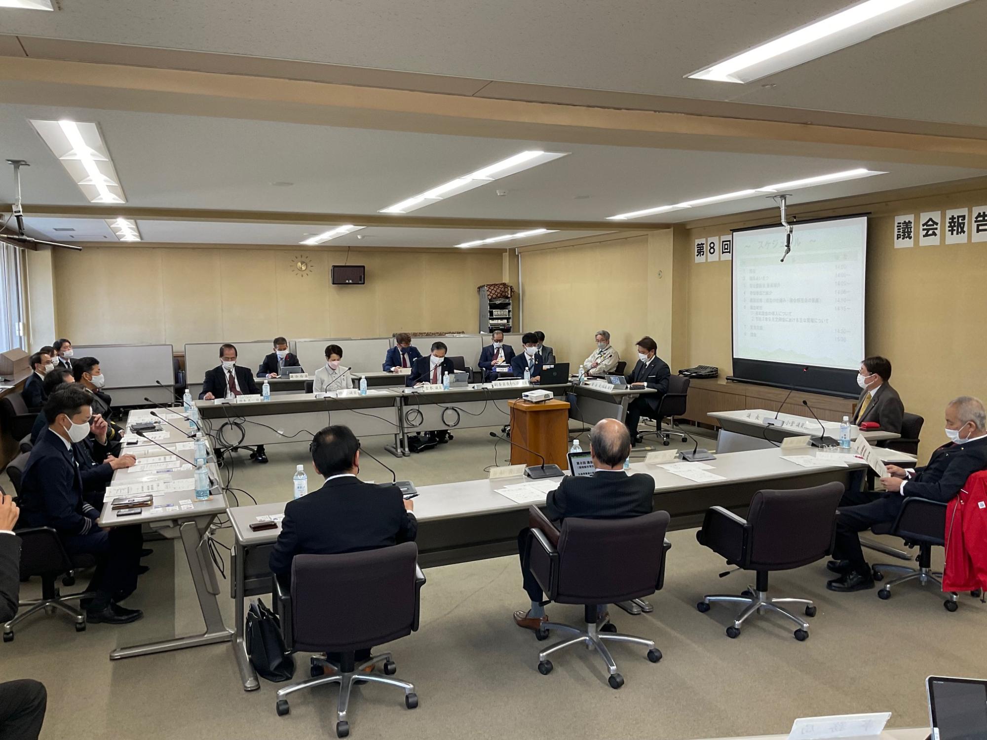 会議室で、多数の参加者がコの字型に並べられた机に座り、プロジェクターに資料が映し出された報告会の様子の写真