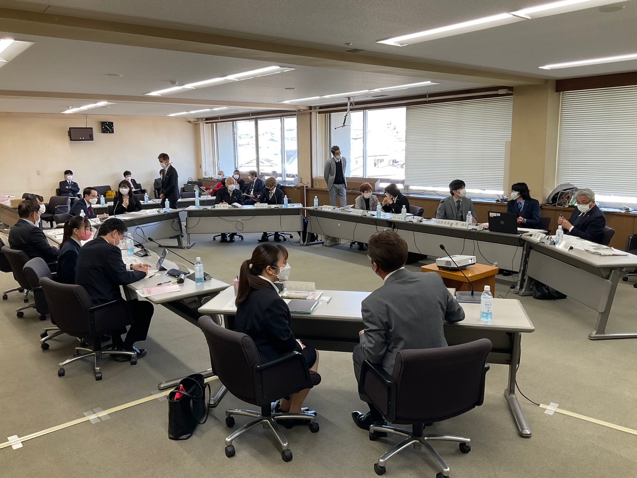 広々とした会議室で、大勢の参加者が楕円型に配置された会議机を囲み、全員マスクを着用して真剣な表情で会議に臨んでいる様子の写真