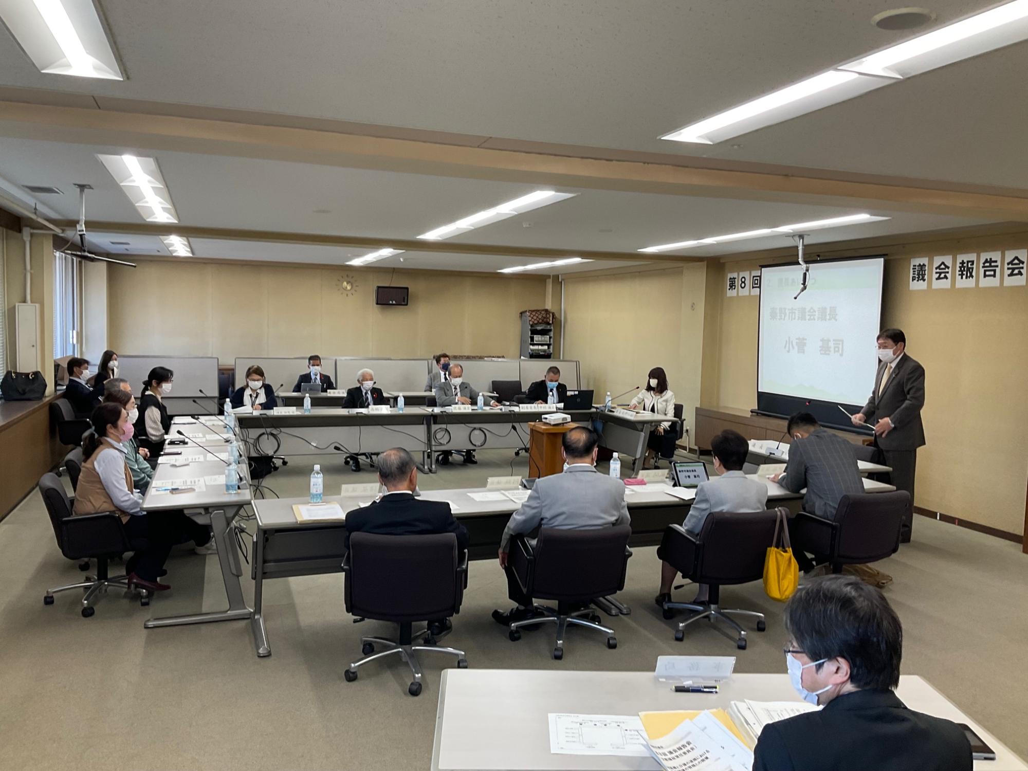 広い会議室で多数の男女がマスクを着用し、コの字型に配置された机を囲んで席に着き、プロジェクターの横で男性が立って報告している様子の写真