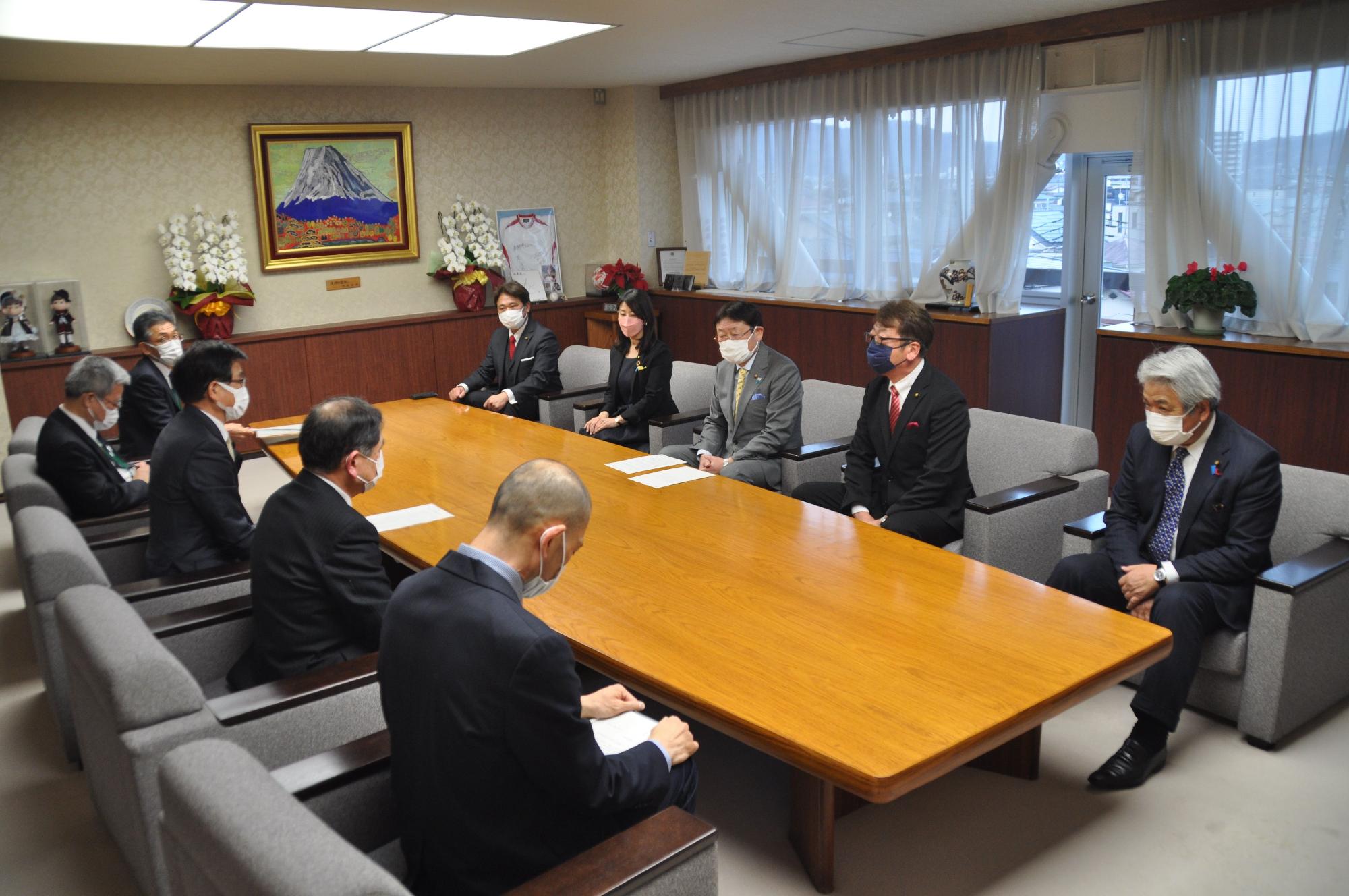 スーツ姿の市長と関係者がマスクを着け左右に分かれて木製の長机を囲み、真剣な面持ちで会談に臨んでいる様子の写真