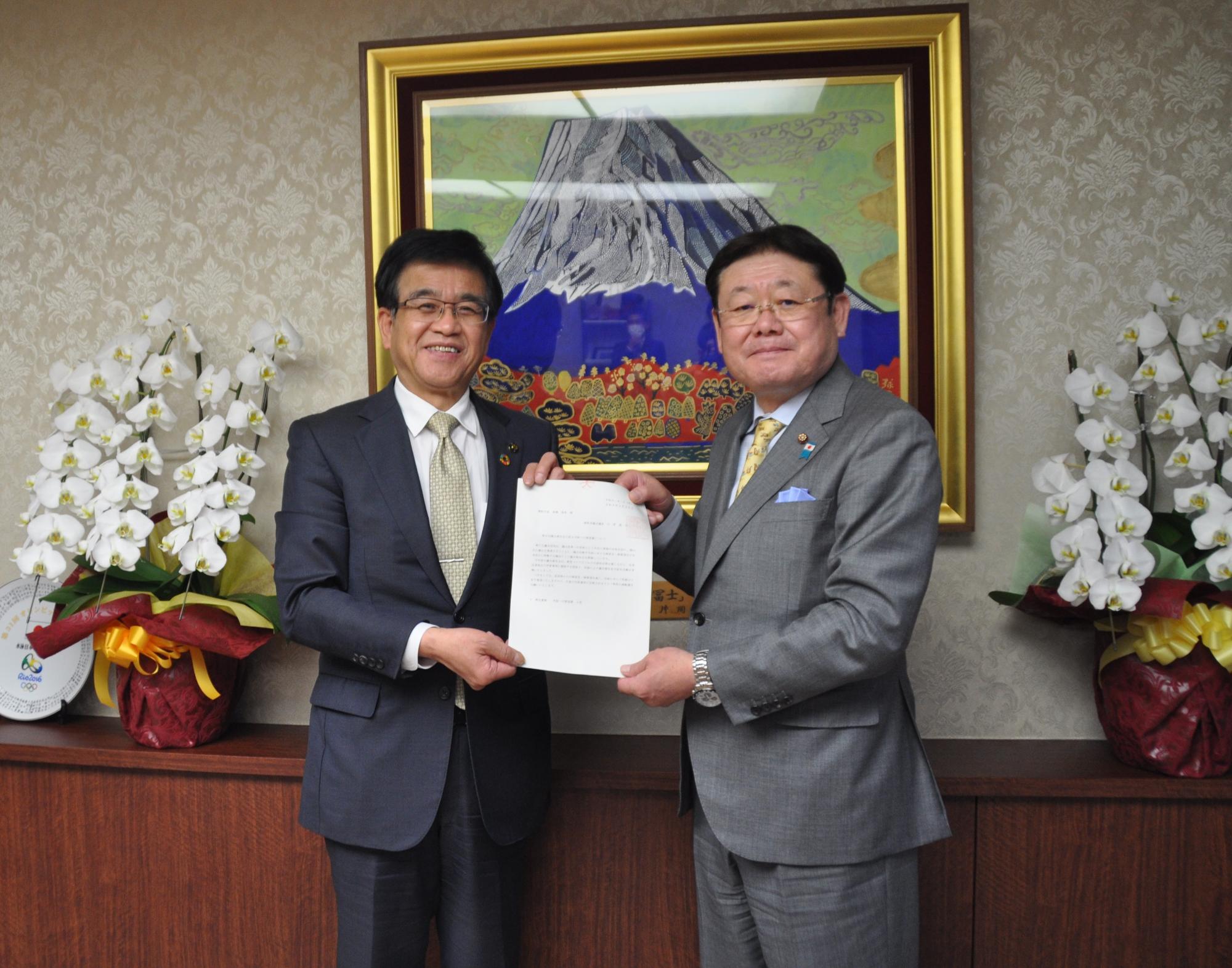 富士山の絵画と豪華な胡蝶蘭の鉢植えを背に、スーツ姿の市長と男性が中央で書類を一緒に持ち、笑みを浮かべながら記念撮影をしている様子の写真