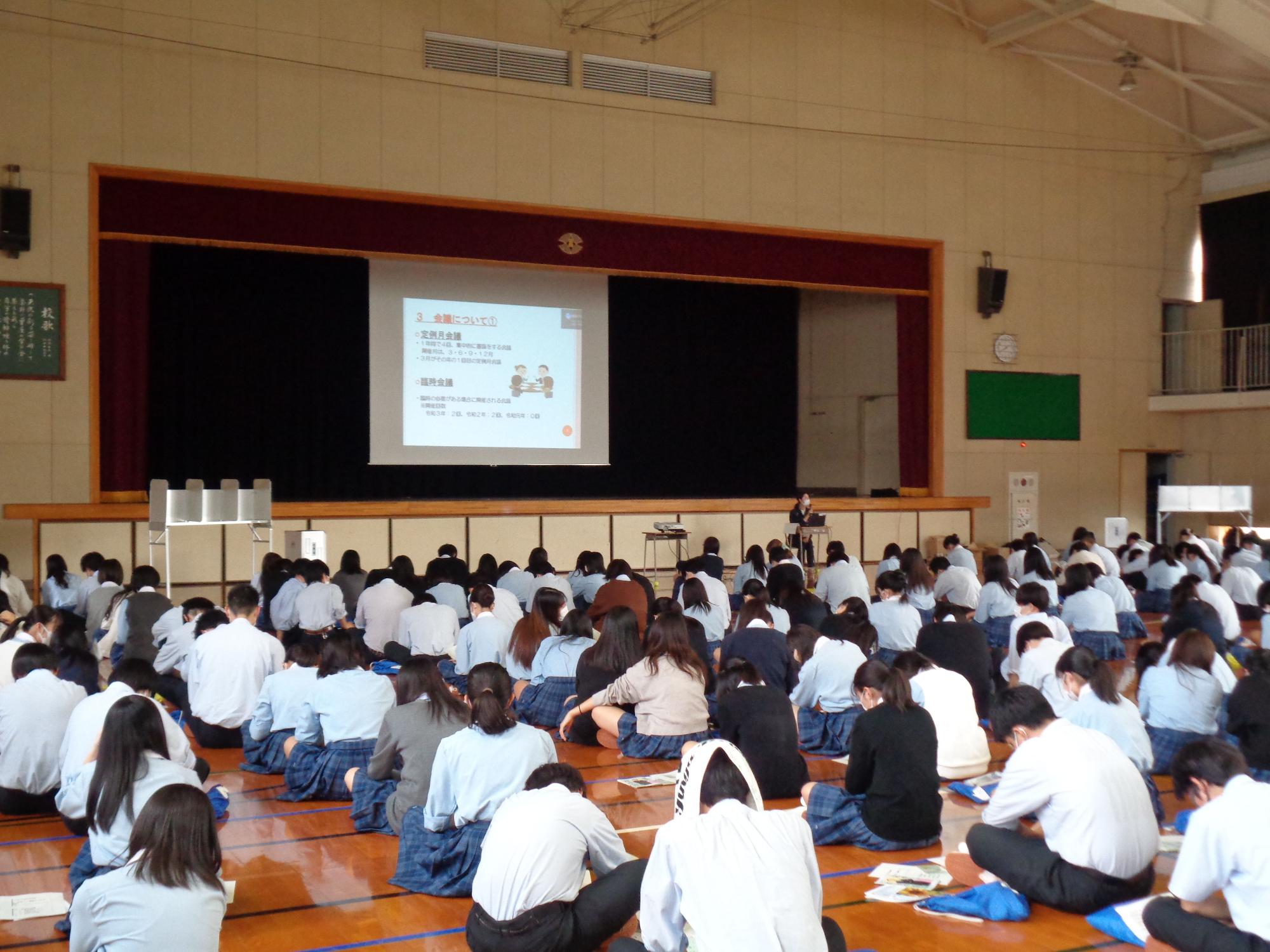 体育館の床に制服姿の生徒が大勢座って壇上を見ており、中央のスクリーンには資料と人物のイラストが投影され、一人の講師が生徒たちの方を向いて座って出前講座が行われている様子の写真